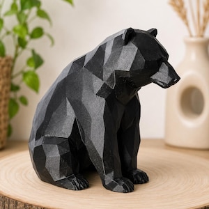 Könnte beinhalten: Eine schwarze, geometrische Bärenskulptur auf einer Holzscheibe. Der Bär besteht aus kantigen Formen, was ihm ein modernes, künstlerisches Aussehen verleiht. Ein dekoratives Element für die Inneneinrichtung.