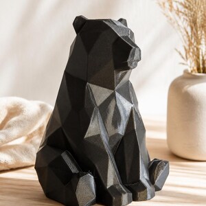 Peut inclure: Une figurine d'ours noir présentant un design géométrique à facettes avec des angles vifs et des surfaces planes. La sculpture est réalisée dans une finition mate sombre et unie, représentant l'animal en position assise, la tête légèrement tournée.