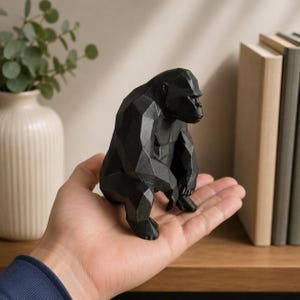 Può includere: Una scultura di gorilla geometrica nera. Il gorilla è seduto con le mani sulle ginocchia. La scultura ha un design sfaccettato ed è realizzata in un materiale scuro. La scultura è su una mensola di legno.