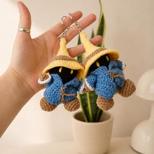 May include: Two crochet keychains featuring figures with black faces, yellow pointed hats, and blue coats with brown accents. Each figure has brown hands and feet, and is attached to a silver metal keyring with a short chain for easy attachment.