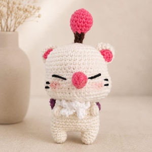 May include: A cream-colored crochet plush toy featuring a round head with a pink pom-pom antenna, pink inner ears, and a pink nose. The figure has black embroidered eyes, small whiskers, a white neck ruffle, and small purple wings on its back.