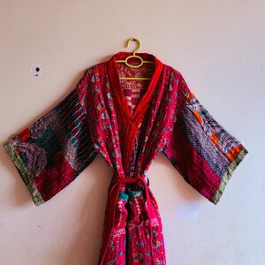May include: A long-sleeved robe featuring a vibrant patchwork design in shades of red, pink, and orange. The garment includes a matching fabric tie belt at the waist, wide sleeves, and intricate kantha-style stitching throughout the fabric.