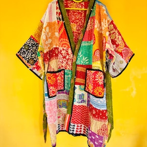 May include: A patchwork kimono-style jacket with a vibrant mix of colors and patterns. The jacket features a variety of fabric patches in red, orange, green, and purple, with black trim and pockets. It hangs on a yellow hanger against a yellow background.