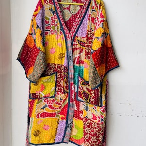 Op de afbeelding: Een patchwork kimono-jas in levendige kleuren, waaronder rood, geel en paars. De jas heeft een V-hals, zakken en een strikceintuur. De stof is bedrukt met bloemen- en geometrische patronen en hangt aan een gele hanger.