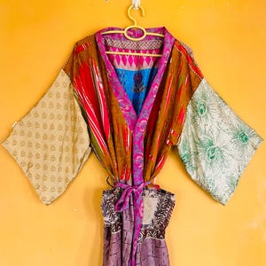 May include: A long-sleeved robe featuring a patchwork design with various patterns including peacock feathers, geometric shapes, and floral motifs in shades of brown, pink, blue, and cream. The garment includes a matching fabric tie belt.