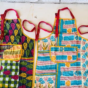 May include: A collection of colorful aprons with unique patterns. The aprons feature a variety of designs, including floral, leaf, and geometric patterns. Each apron has a pocket and red straps. The aprons are made of a patchwork fabric with yellow trim.