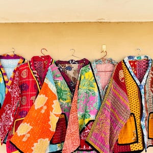 May include: Five colorful, patchwork jackets hanging on a wall. Each jacket features a unique combination of patterns and colors, including blue, red, orange, pink, and yellow. The jackets are made of a quilted fabric and have a relaxed, open front design.