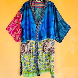 May include: A colorful kimono robe with a patchwork design. The robe features a mix of patterns and colors, including blue, pink, green, and gold. It has long sleeves and pockets. The robe is hanging on a yellow hanger against a yellow background.