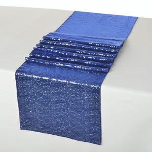 Puede incluir: Un camino de mesa de lentejuelas azul real. El camino de mesa rectangular está drapeado sobre una superficie blanca. Las lentejuelas crean un efecto texturizado y brillante. El camino de mesa está diseñado para uso decorativo.