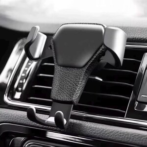 May include: A black car phone mount featuring a textured, leather-like central panel and two side-gripping arms. The device includes a bottom support bracket with two small hooks and is designed to attach securely to a vehicle air vent slat.