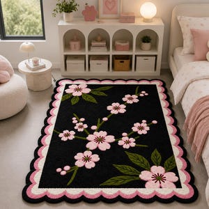 May include: A rectangular area rug featuring a black background with a central cherry blossom branch design in shades of pink and green. The rug has a decorative scalloped border in alternating layers of white and pink, creating a distinct frame.