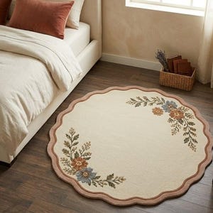 May include: A round, cream-colored rug with a scalloped edge and floral design. The rug features a border in a muted rose color and floral accents in shades of blue, orange, and green. The rug is placed on a dark wood floor.
