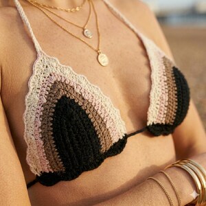 May include: A crochet bikini top featuring a triangular design with horizontal stripes in cream, light pink, brown, and black. The edges of the cups have a scalloped finish, and the garment includes thin crochet straps that tie around the neck.