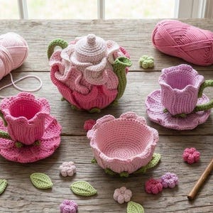 Könnte beinhalten: Ein Häkel-Teeservice bestehend aus einer Teekanne, zwei Tassen mit Untertassen und einer kleinen Schale, gestaltet mit rosa und hellrosa Blütenblattmotiven und grünen Henkeln. Zwei Knäuel rosa Garn und eine Häkelnadel aus Holz liegen dabei.
