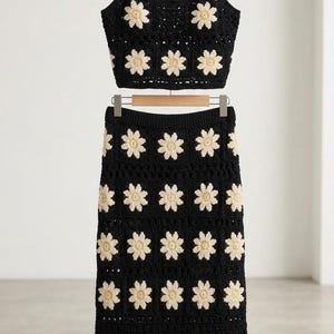 May include: A two-piece crochet set consisting of a black cropped tank top with thin straps and a matching midi-length skirt. Both pieces feature a repeating pattern of cream-colored daisy motifs against a solid black background.