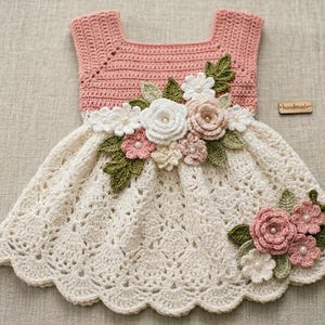May include: A handmade crochet dress in pink and cream, adorned with floral embellishments. The bodice is a solid pink, while the skirt is cream-colored with a lacy pattern. The dress features a square neckline and is decorated with white, pink, and green crochet flowers.