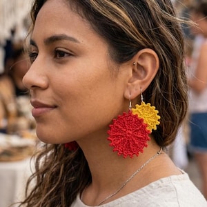 Puede incluir: Pendientes de ganchillo rojos y amarillos con diseño floral. Los pendientes están hechos de hilo y tienen forma circular con un patrón texturizado. Los pendientes cuelgan de ganchos plateados.