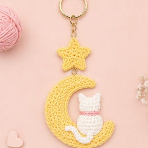 May include: A crochet keychain featuring a yellow star attached to a gold-tone metal ring, connected to a yellow crescent moon. A small white cat with a pink collar is stitched onto the center of the moon, with its tail curved along the bottom edge.