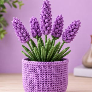 Könnte beinhalten: Eine handgefertigte gehäkelte Lavendelpflanze in einem lila Topf. Die Lavendelblüten sind leuchtend lila, mit grünen Stielen und Blättern. Der Topf ist passend lila, mit einem strukturierten, gewebten Aussehen. Die Anordnung ist vor einem weichen lila Hintergrund.