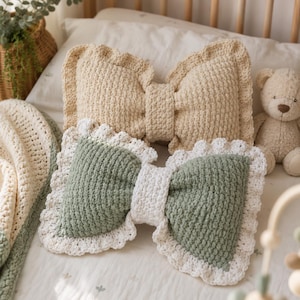 May include: Two bow-shaped throw pillows made of crocheted yarn. One pillow is solid beige with a ruffled edge, and the other is sage green with a white centre knot and a white scalloped crochet border. Both pillows feature a textured stitch pattern.