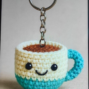 May include: A crocheted coffee cup keychain with a smiling face. The cup is cream, teal, and brown, with a silver chain and keyring. The keychain is a handmade item.