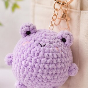 May include: A round, light purple crochet frog keychain featuring a textured yarn surface, small protruding limbs, and black embroidered eyes. The plush figure is attached to a gold-toned metal split ring and a lobster clasp for easy attachment.