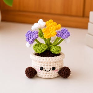 May include: A crocheted flower pot with a smiling face, brown feet, and colorful flowers. The flowers are white, yellow, and purple. The pot is beige and filled with brown yarn to resemble soil. A charming handmade decorative item.
