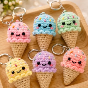 May include: Six crocheted ice cream cone keychains in various pastel colors including pink, blue, mint green, yellow, purple, and peach. Each keychain features a tan cone, a smiling face with black bead eyes, and colorful embroidered sprinkles.