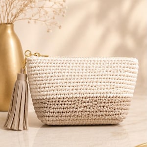 Puede incluir: Un pequeño estuche rectangular de ganchillo con un diseño degradado de dos tonos que va del crema en la parte superior al beige claro en la base. El estuche incluye una cremallera dorada adornada con una borla de piel sintética beige claro.