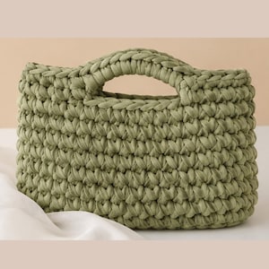 May include: A sage green handbag crafted from thick, chunky yarn in a dense crochet stitch pattern. The bag features a rectangular body with a rounded top edge and two integrated, circular cut-out handles formed by the continuous crochet work.