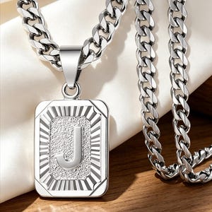 May include: A silver-toned metal necklace featuring a rectangular pendant with a textured border and a polished letter J in the center. The pendant hangs from a thick, interlocking curb chain with a high-shine, reflective metallic finish.
