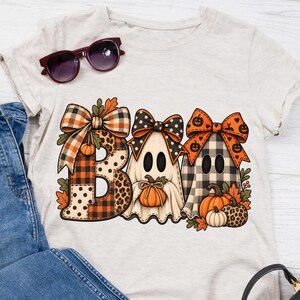 May include: A heathered beige t-shirt featuring a graphic print of the word BOO, where the letters are stylized as ghosts and pumpkins. The design includes orange, black, and white plaid, leopard print, and polka dot patterns with bows.