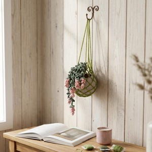 Puede incluir: Un macetero de macramé colgante con una planta colgante verde y rosa. El macetero está suspendido de un gancho decorativo en una pared de paneles de madera clara. Un libro, una pequeña taza rosa y carretes de hilo están sobre una mesa de madera.