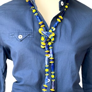 Puede incluir: Un collar largo con cuentas de vidrio azul y amarillo. El collar se lleva sobre una camisa azul.