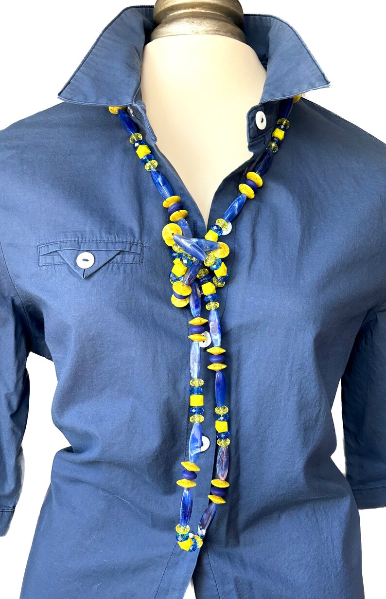 Puede incluir: Un collar largo con cuentas de vidrio azul y amarillo. El collar se lleva sobre una camisa azul.