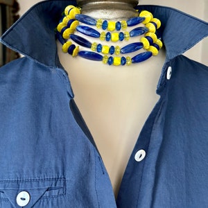 Puede incluir: Un collar de varias capas con cuentas azules y amarillas. El collar est&aacute; hecho de cuentas de diferentes formas y tama&ntilde;os, incluyendo cuentas redondas, ovaladas y cuadradas. El collar se lleva sobre un maniqu&iacute; que lleva una camisa azul.
