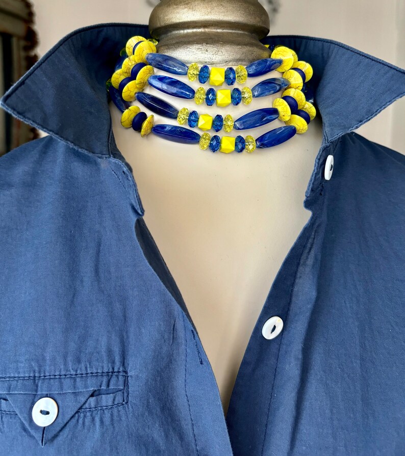 Puede incluir: Un collar de varias capas con cuentas azules y amarillas. El collar est&aacute; hecho de cuentas de diferentes formas y tama&ntilde;os, incluyendo cuentas redondas, ovaladas y cuadradas. El collar se lleva sobre un maniqu&iacute; que lleva una camisa azul.