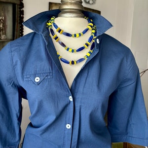 Puede incluir: Un collar de varias capas con cuentas de vidrio azul y amarillo. El collar est&aacute; sobre una camisa azul.