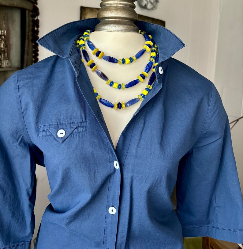 Puede incluir: Un collar de varias capas con cuentas de vidrio azul y amarillo. El collar est&aacute; sobre una camisa azul.