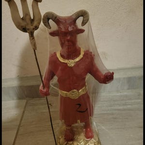 Op de afbeelding: Een rood gehoornd beeldje op een gouden voet, verpakt in doorzichtig plastic. Het figuur draagt een gouden ketting en riem, houdt een hoge drietand vast en heeft het cijfer 2 met zwarte inkt op de voorkant van het rode gewaad geschreven.