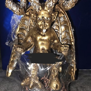 May include: A gold-colored figurine of a horned creature with large wings, pointed ears, and a small goatee. The figure has a muscular torso, clawed feet, and is partially wrapped in clear plastic packaging with a black bar obscuring the midsection.