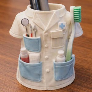 Puede incluir: Un original porta cepillos de dientes con forma de bata de médico, con bolsillos azul claro y un emblema de cruz. Contiene instrumentos dentales, pasta de dientes y un cepillo de dientes con cerdas verdes. El soporte está sobre una superficie de madera.