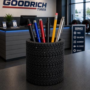 May include: A black tire-shaped pencil holder filled with pens and a pencil. The pens are black, blue, red, and silver. The pencil is yellow with a pink eraser. The background features the Goodrich Tires logo and a service sign.