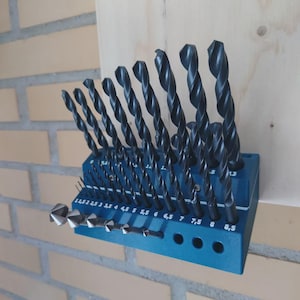 Può includere: Un set di punte da trapano nere in un supporto blu. Le punte sono disposte per dimensione, con misure in millimetri stampate sul supporto. Lo sfondo include un muro di mattoni e una superficie in legno.