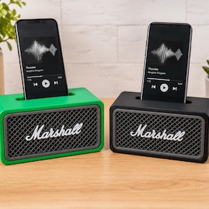 Puede incluir: Dos bases de carga para altavoces de la marca Marshall, una verde y otra negra, cada una con una rejilla frontal de malla gris y una ranura superior que sostiene un teléfono inteligente con una onda sonora y el texto Thunder en pantalla.
