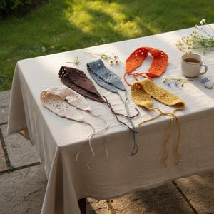 May include: A collection of handmade crochet headbands in various colors, including cream, brown, blue, orange, and yellow, are displayed on a table. Each headband features a unique openwork design and is tied with a matching string.