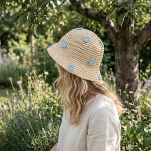 Może przedstawiać: Beżowy szydełkowy kapelusz typu bucket hat z małymi, jasnoniebieskimi pięciopłatkowymi kwiatkami rozmieszczonymi na koronie i rondzie. Kapelusz wykonany jest z plecionego materiału przypominającego słomę o teksturalnym, powtarzalnym splocie.
