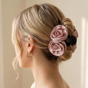 Könnte beinhalten: Eine schwarze Haarklammer aus Kunststoff, verziert mit zwei großen, zartrosa Stoffrosen. Das Accessoire ist in einem blonden Dutt befestigt und zeichnet sich durch eine glatte, matte Oberfläche der Blütenblätter und eine glänzende Klammer aus.