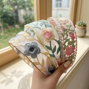 May include: A coiled length of sheer white mesh ribbon featuring intricate floral embroidery. The design includes delicate green vines, soft pink blossoms, and a prominent grey flower, all stitched with fine thread onto a translucent fabric base.