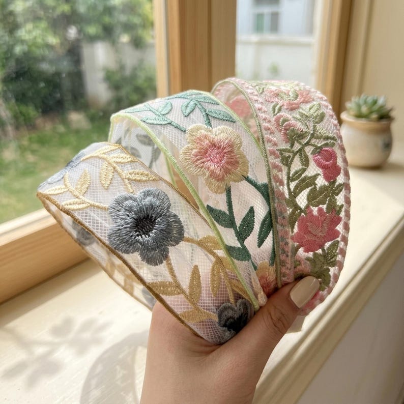 May include: A coiled length of sheer white mesh ribbon featuring intricate floral embroidery. The design includes delicate green vines, soft pink blossoms, and a prominent grey flower, all stitched with fine thread onto a translucent fabric base.